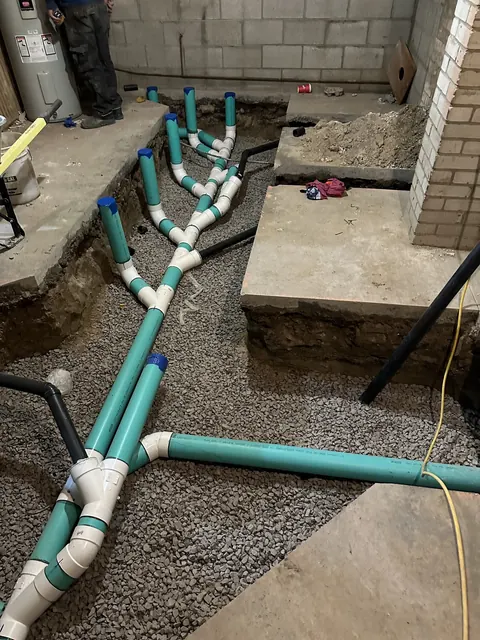 Green drain pipes and white fittings laid in gravel before a basement floor is poured.